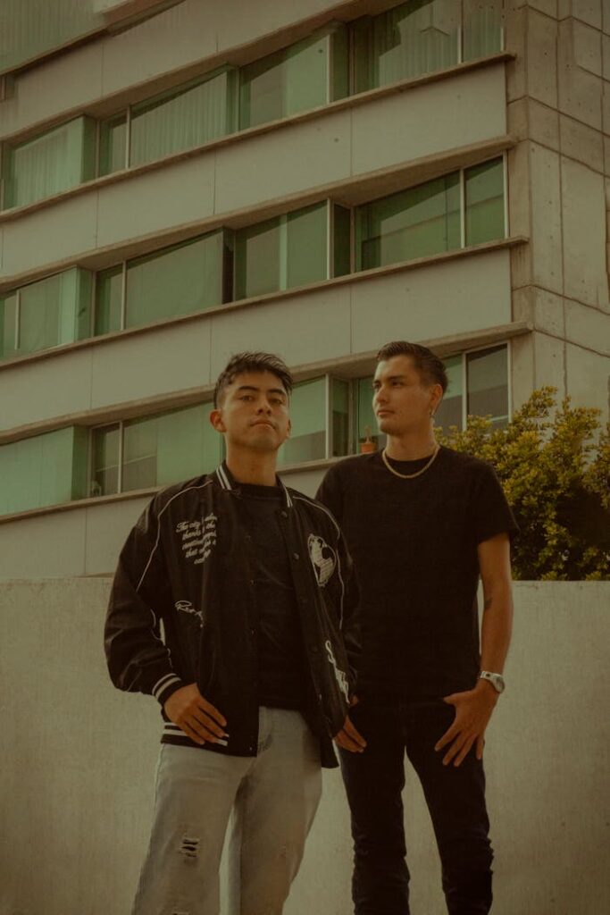 Two young men pose confidently in front of a modern building, showcasing urban street style.